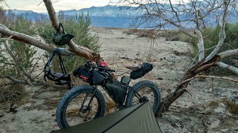 Left side view of a Surly fat bike standing in a sandy field, in front of a small tree, with mountains in the background