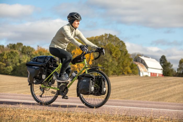 Cyclist rides a Surly Disc Trucker bike on the shoulder of a country road while passing a picked field and barn