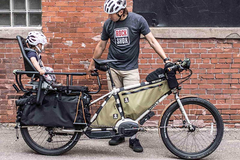 Cyclist stands on the left side of  a Surly Big Easy bike while looking back at a child seated in the rear carrier mount