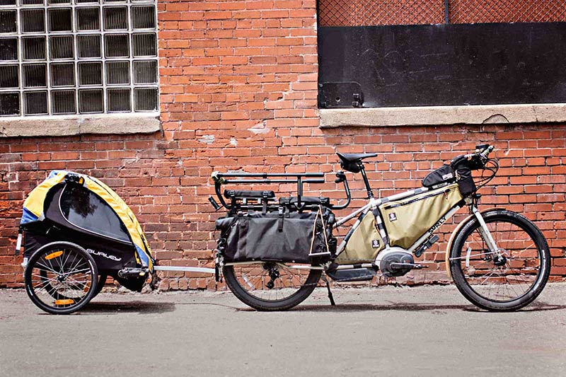 Right view of a white Surly Big Easy bike with gear and a Burley trailer attached, on concrete next to a brick building