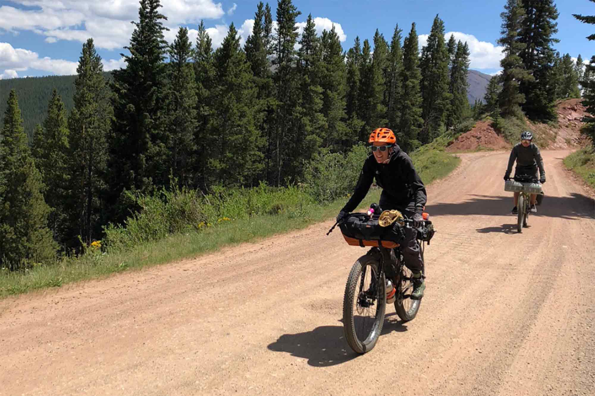 Cyclist smiles riding up a hill on a gravel road in the pine trees to the side with another rider tailing behind