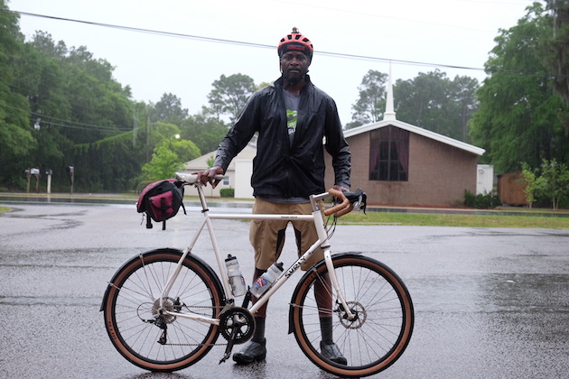 Cyclist standing in the rain behind a Surly Midnight Special bike, white, with a seat pack, right profile view