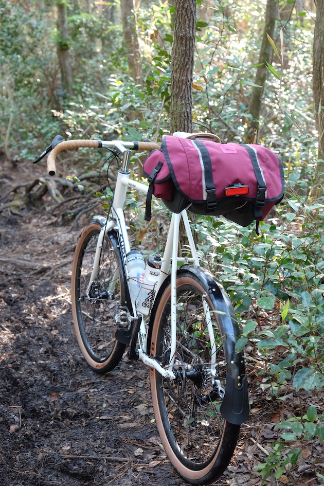 Surly Midnight Special bike, white, with red seat pack facing away down a dirt trail in the woods