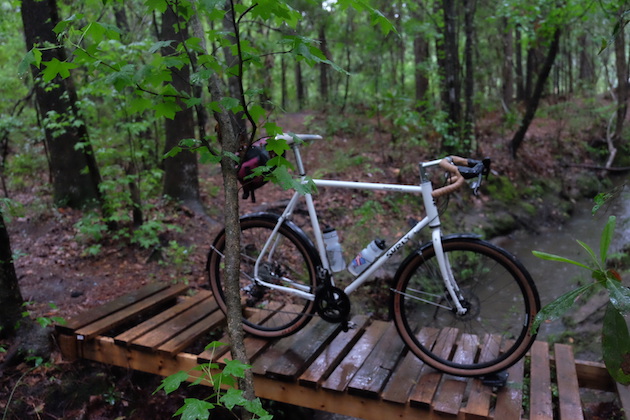 Right side view of a Surly Midnight Special bike, white, with seat pack, stand on a wet, wood double pallet in the trees