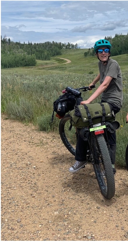 Cyclist on the side of a gravel road on a black Surly ECR bike loaded with gear in a hilly prairie with trees behind