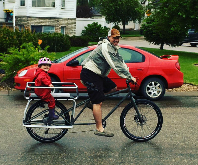 Right side view of a cyclist riding a Surly Big Dummy bike with a child in back, on a street with a car in background