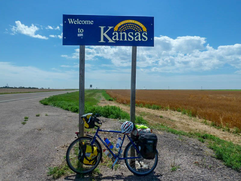 Left side view of a blue Surly bike loaded with gear, on the side of a roadway, under a Welcome to Kansan sign