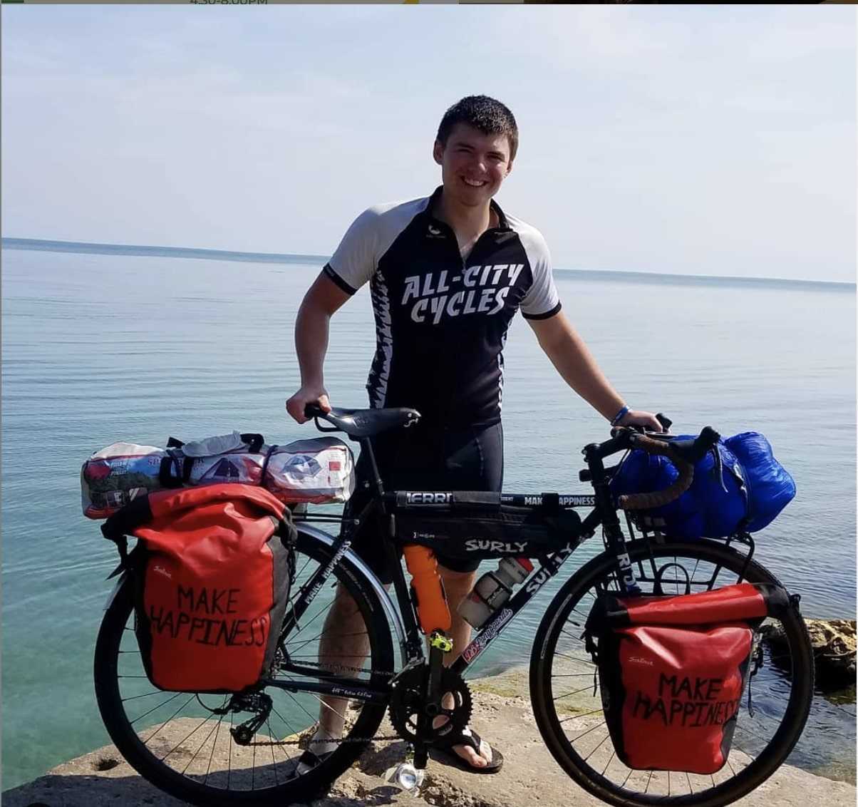 Right profile view of a black Surly bike with gear, and rider standing behind, on a flat rock at the edge of a lake