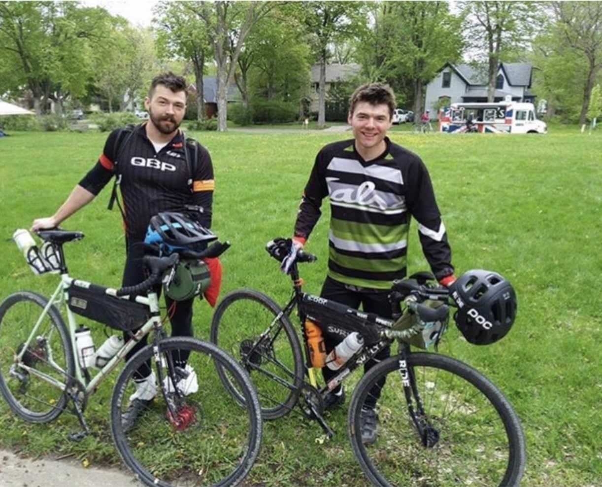 Two cyclists pose while standing on the edge of a grass field with a gray house, a white van and trees in the distance