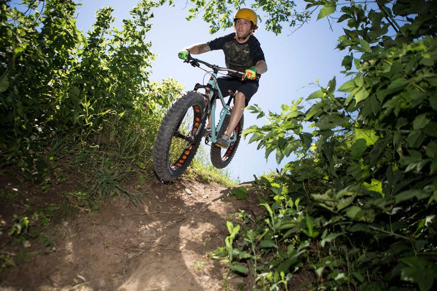 Upward, front view of a cyclist, on a light blue Surly Wednesday fat bike, riding down a hill, on a dirt trail