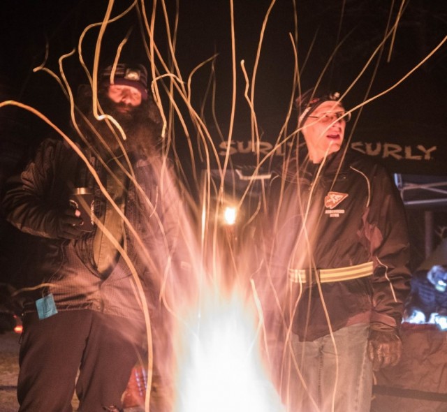 A bright fireworks flame at night, with two people standing behind, and Surly canopies in the background