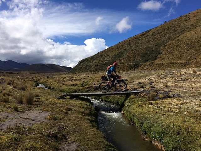 Right side view of a cyclist riding a Surly Pugsley bike across a narrow bridge, over a stream, in a grassy field