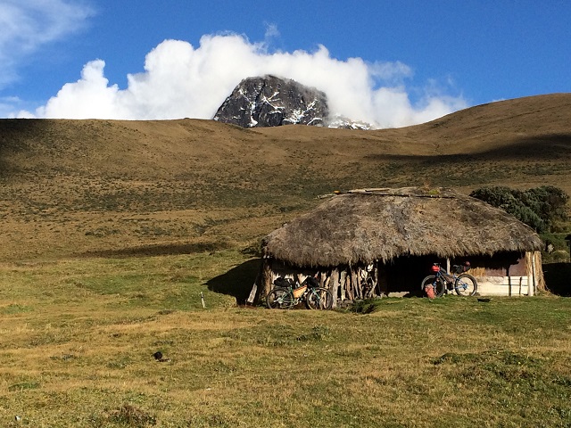 Two Surly bikes in front of a thatch roof home in a field, with a mountain in the background