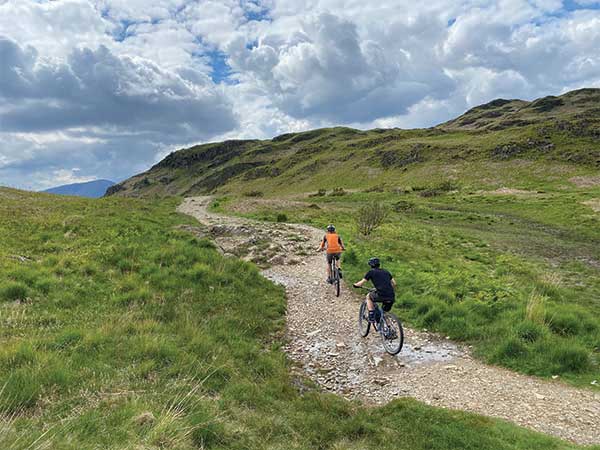 Two cyclists riding up rocky trail along grassy hills