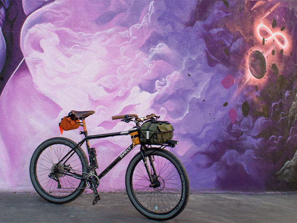 Bridge Club bike with saddle bag and front rack and bag parked next to mural on side of building