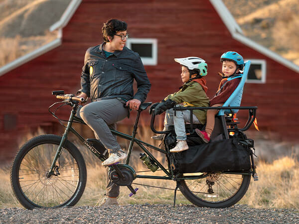 Abner on Big Easy, foot down, looking back at two children sitting on back of bike, outside on gravel, house in background