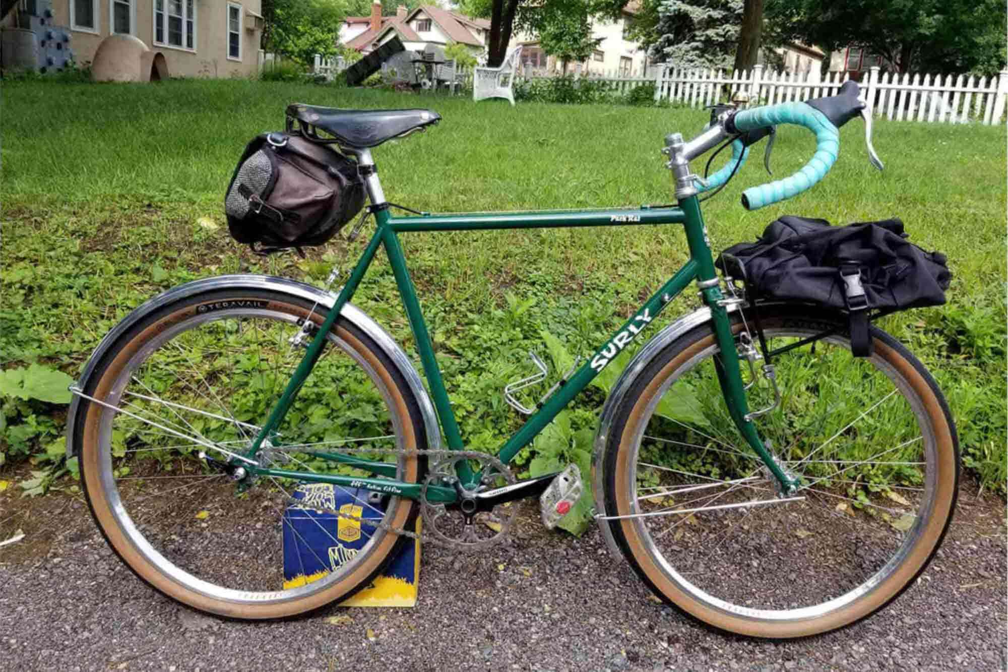 Right side view of a green Surly Pack Rat with gear packs on a sidewalk in front of a yard with a white picket fence