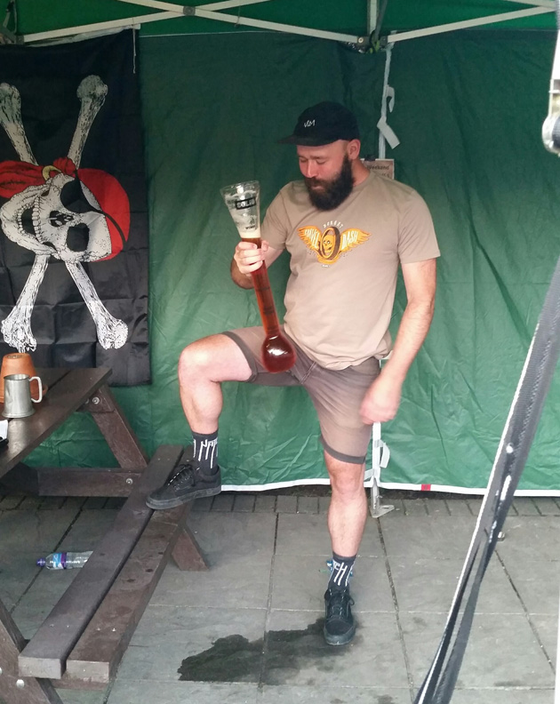 Person looks down at a long narrow glass of beer and  inside of a green pop up canopy on a wood floor