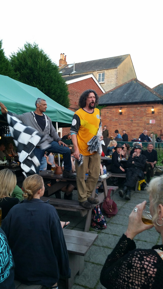 A group gathered at a patio listening to a person in a yellow shirt standing on a picnic table with building behind