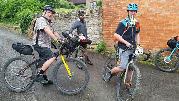 Three cyclist stand with their bikes on a paved hill in front of a orange brick building