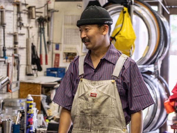 Kaneyan in bike repair shop wearing overalls, tools on wall