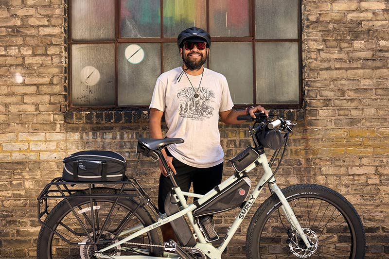 Alexis standing smiling wearing sunglasses and bike helmet with Surly Skid Loader in front of brick building