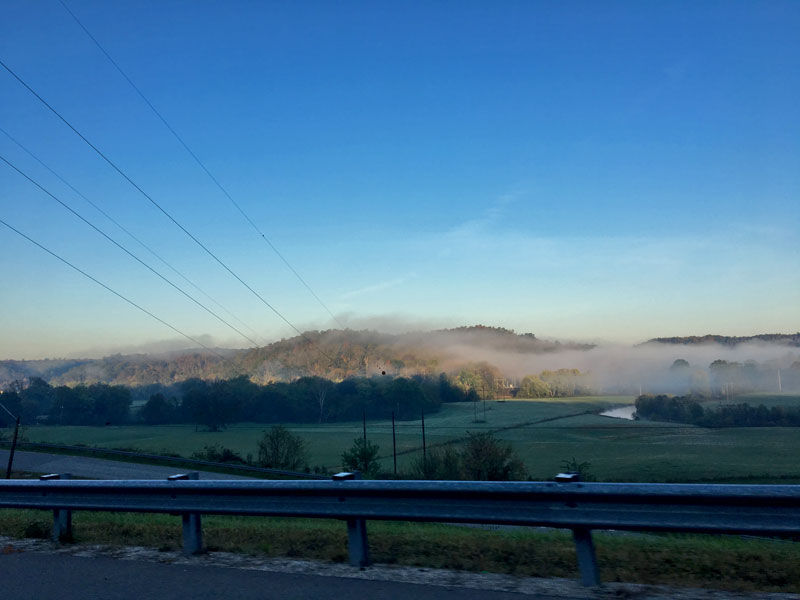 View from  a roadside guardrail with green fields, tree and fog covered hills in the distance