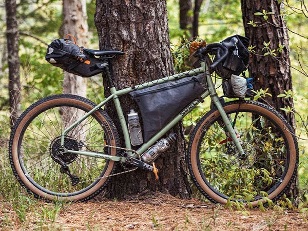 Grappler leaning against tree in forest with seat pack, framepack, handlebar bag, front rack, and two water bottles