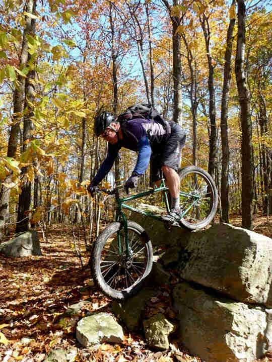 Downward right side rear view of a cyclist riding a Surly Krampus bike, green, down a muddy trail in the woods