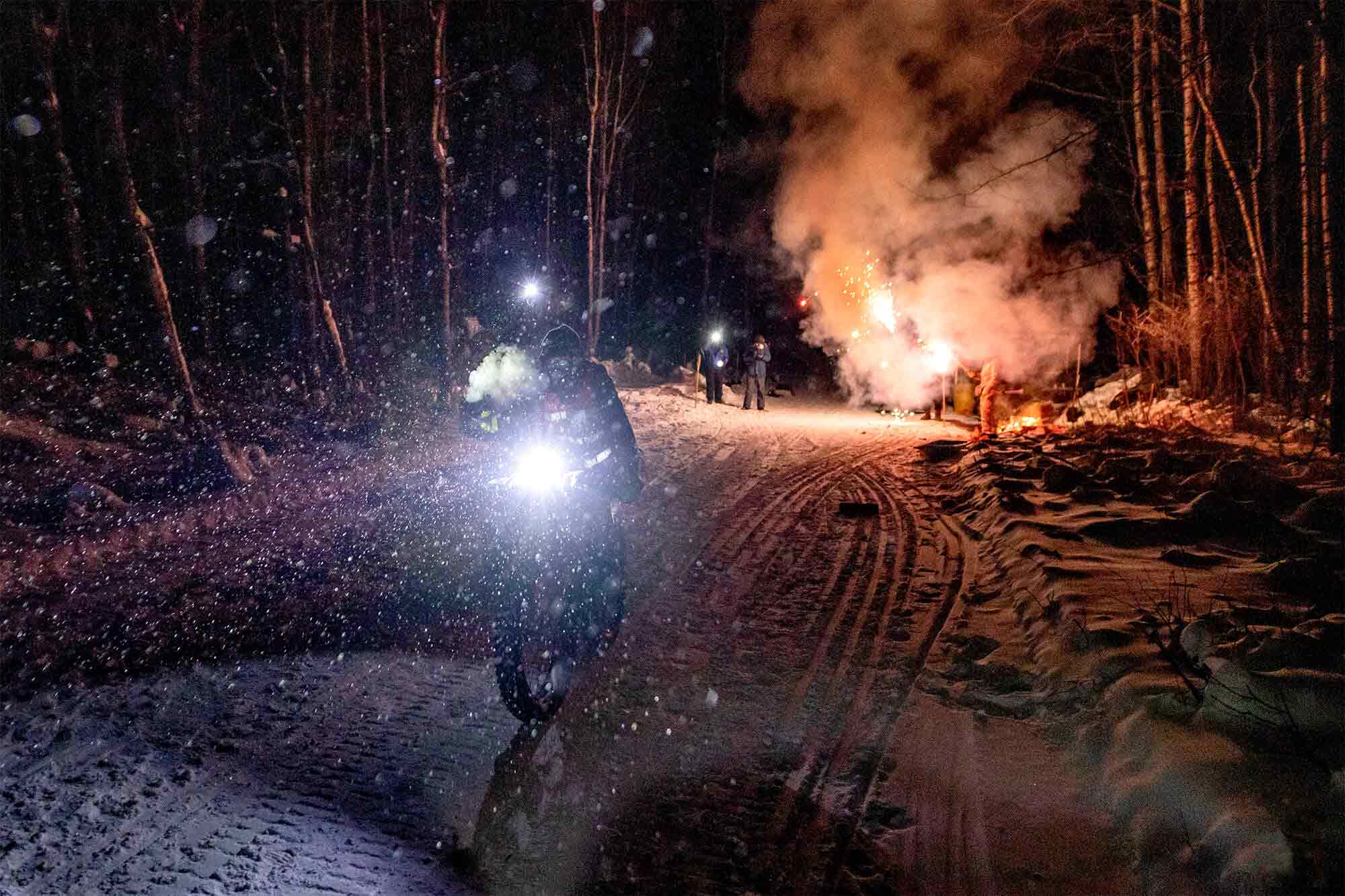 Cyclist riding a bike with a headlight, in the snow at night with fireworks exploding on the ground behind 