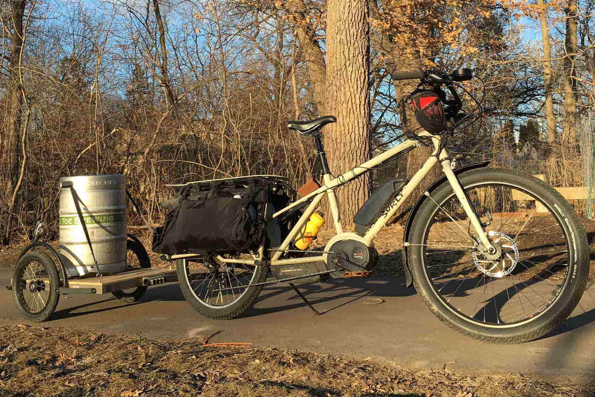 Right profile view of a tan Surly Big Easy bike with gear pulling a trailer with keg on a paved trail with trees behind