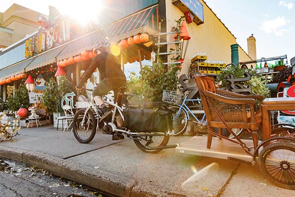 Left side view of a cyclist riding a Surly Big Easy bike with a trailer, past a store front on a sunny day