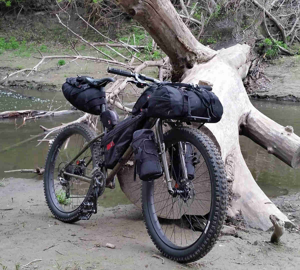 Front, right side view of an olive Surly Instigator bike, parked on a muddy stream shore, in front of a fallen tree