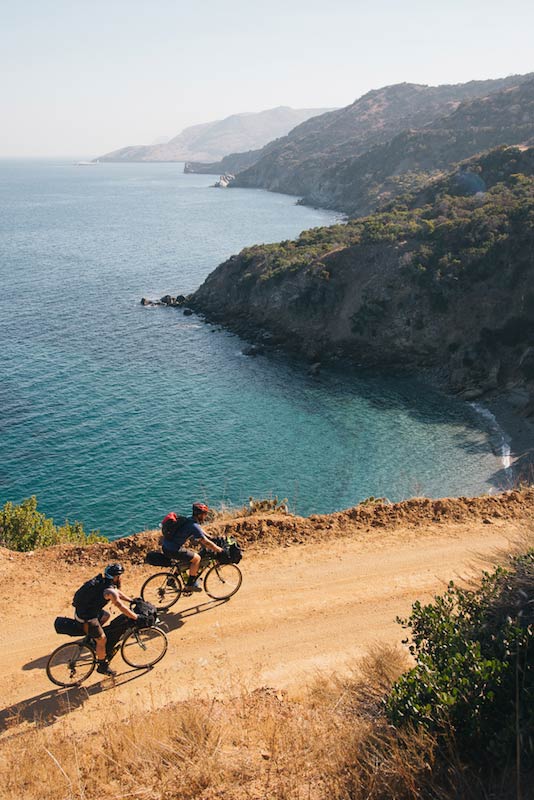 Overhead view of two cyclists with gear loaded bikes, riding down a gravel road on a cliff of a an ocean bay