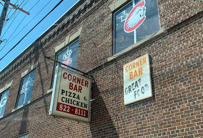 Corner Bar Pizza & Chicken sign on side of brick building with sports team logos in the windows on sunny day