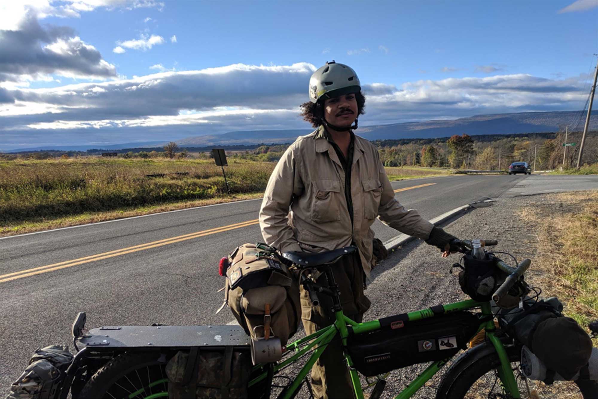 Cyclist standing on a wood bridge over a river with a green Surly Big Dummy bike with blue sky and clouds in the sky