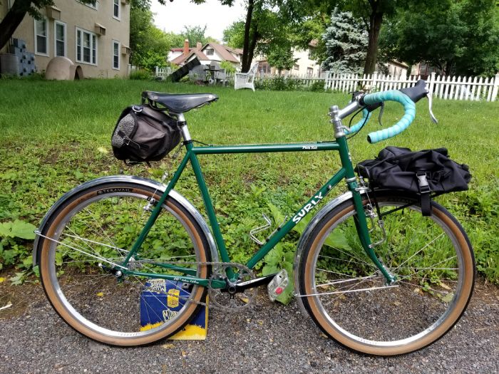 Right side view of a green Surly Pack Rat with gear packs on a sidewalk in front of a yard with a white picket fence