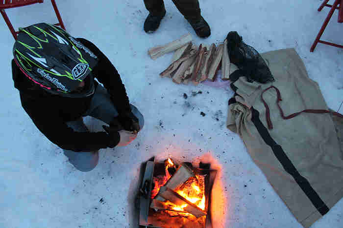 Downward view of a person wearing a helmet, squatting down next to a campfire with kindling next to it