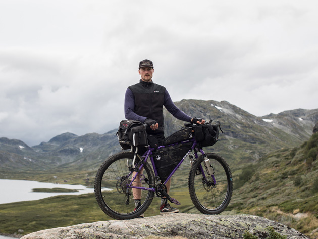 Cyclist standing on a large flat rock on a cloudy day with a lake and mountains in the background