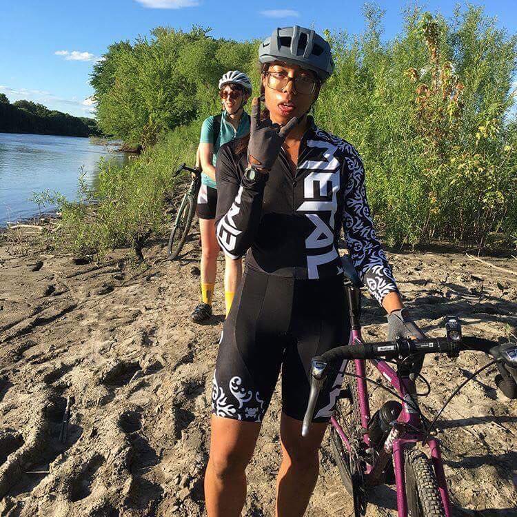 Front view of two cyclists standing inline with their bikes on sandy bank of a river with trees behind, on a sunny day