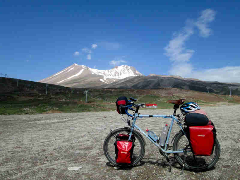 Left side view of a light blue Surly bike, loaded with gear, parked on a rocky field, with mountains in the background