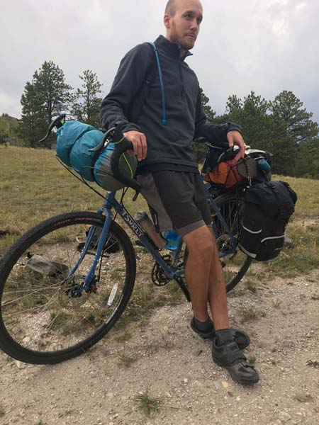 Cyclist on gravel in a field, leaning back on the top tube of a Surly Pacer bike, blue