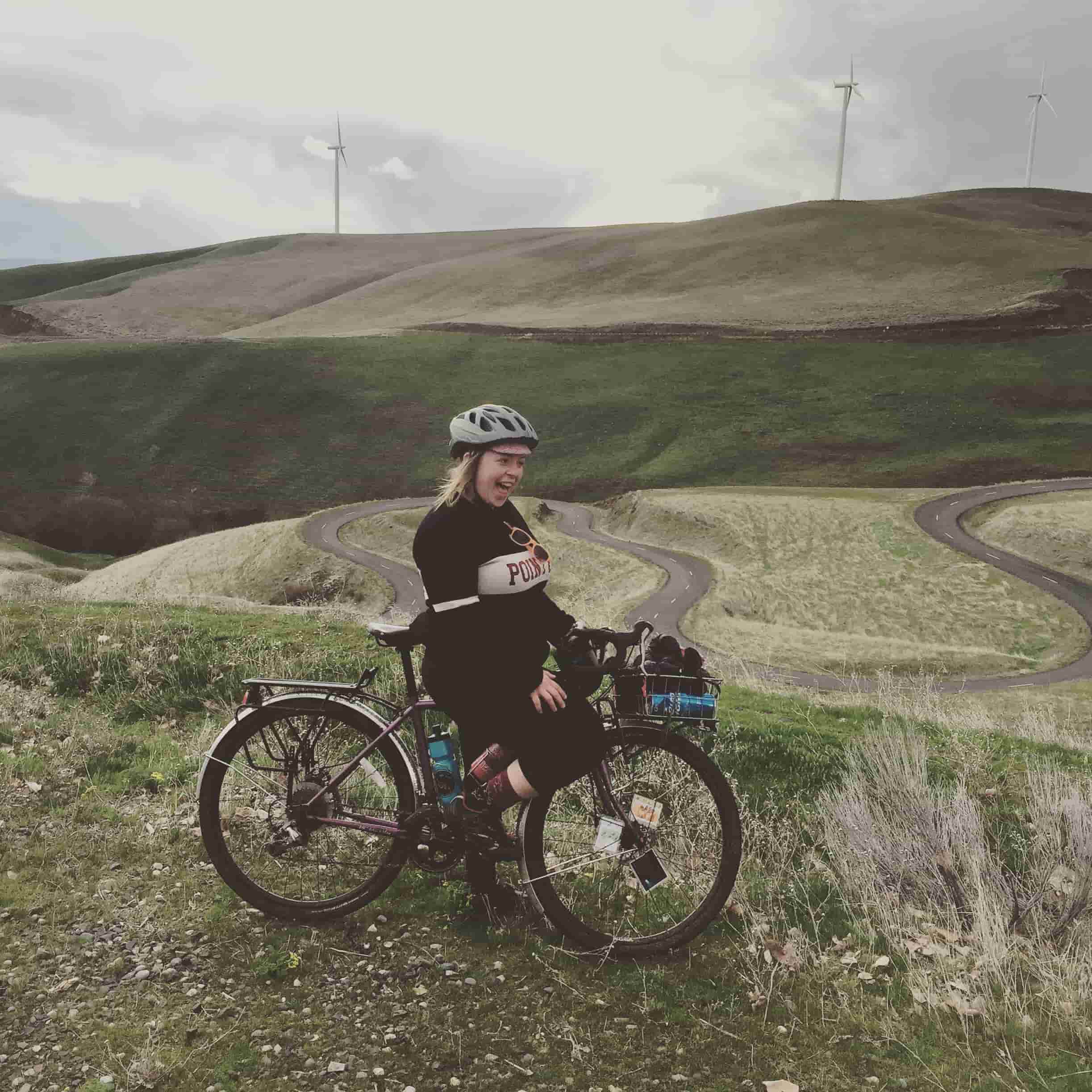 Right side of Cyclist and bike stopped on a grassy patch overlooking a winding trail in rolling hills with windmills