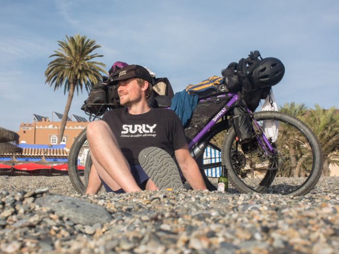 Jack Mac sits with a Surly bike loaded with gear on a gravel beach with palm trees behind