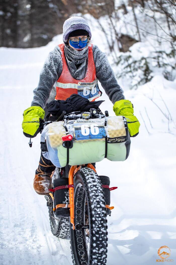 Cyclist wearing winter clothing rides an orange Surly fat bike on a snowy trail in the woods