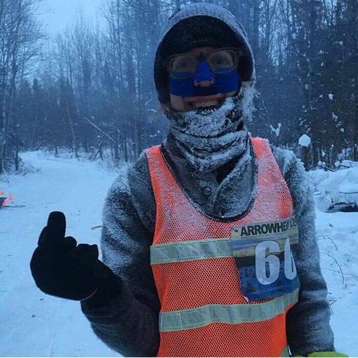 Person wearing winter clothing smiles and sticks up a gloved middle finger on a snowy day in the woods