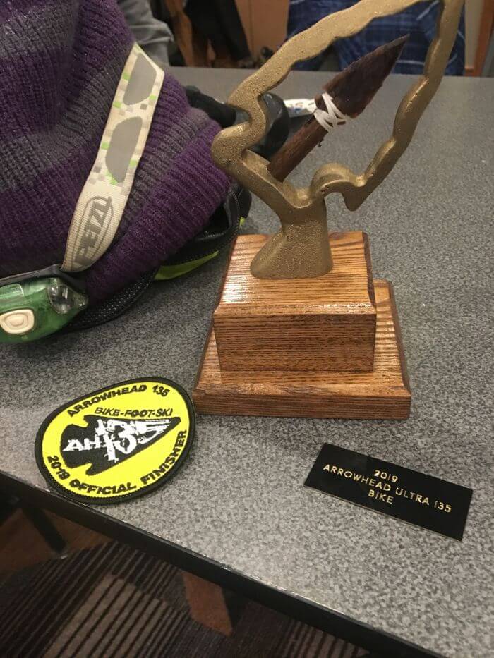 Arrowhead trophy with a wood base, a round shirt patch and a trophy plaque on a table