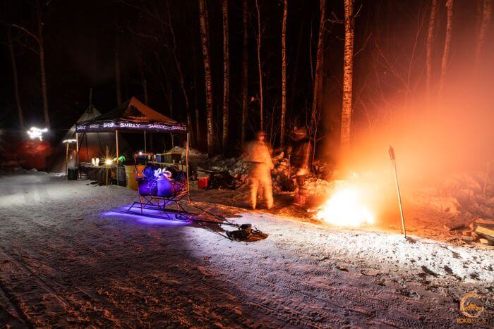 A campfire and a pop up canopy at campsite, in the snowy woods at night