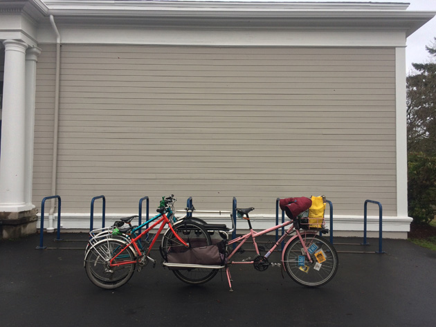 A Surly Big Dummy bike, pink, with two bikes connected to the rear, on pavement in front of a tan building