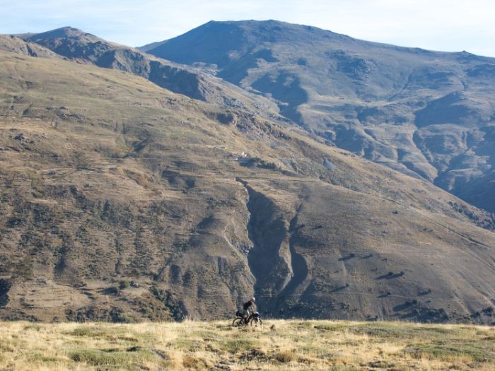 Cyclist with bike loaded with gear rides at the base of grassy mountain range